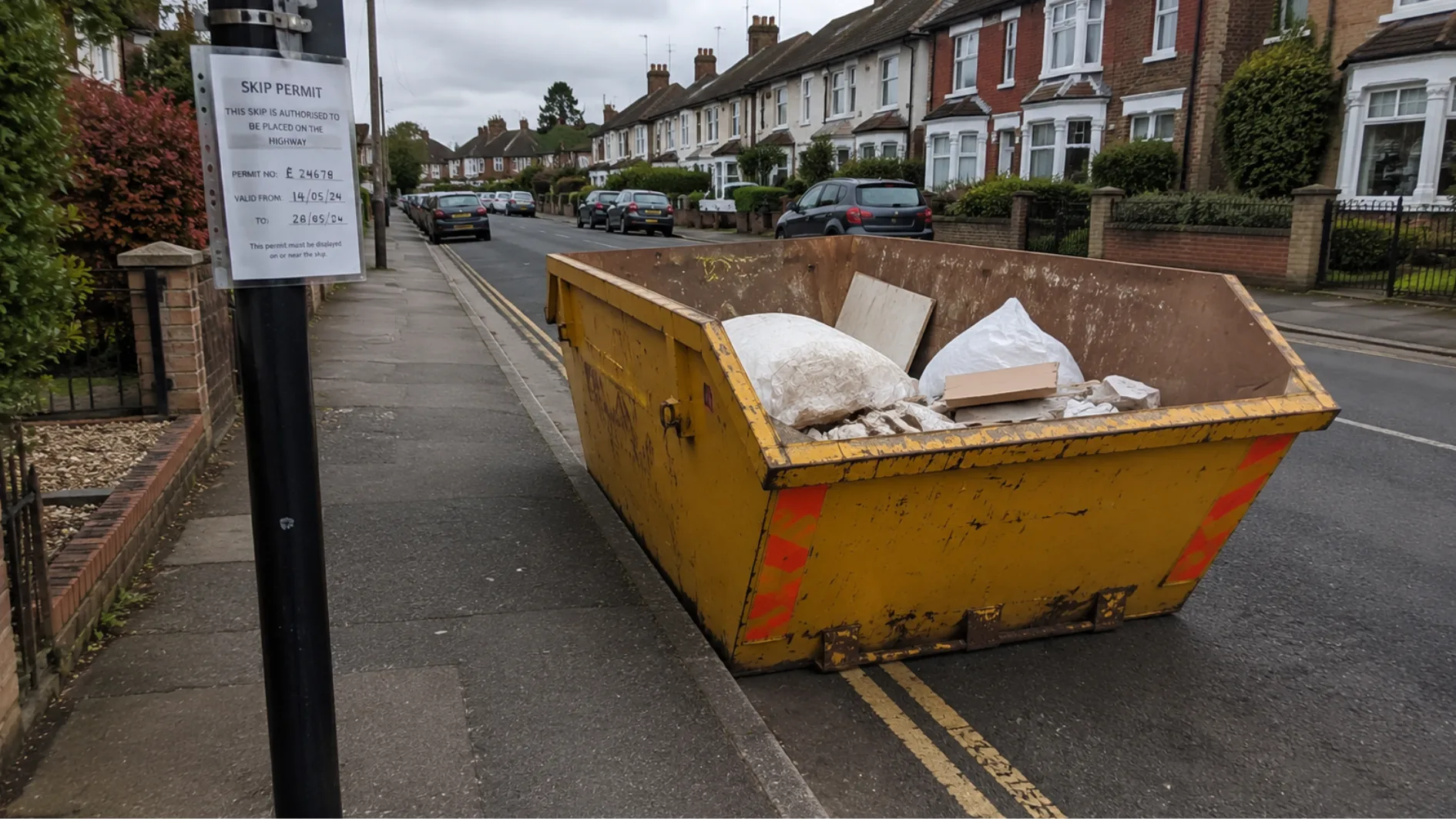 Yellow skip on a residential road beside a pavement with a visible skip permit notice.